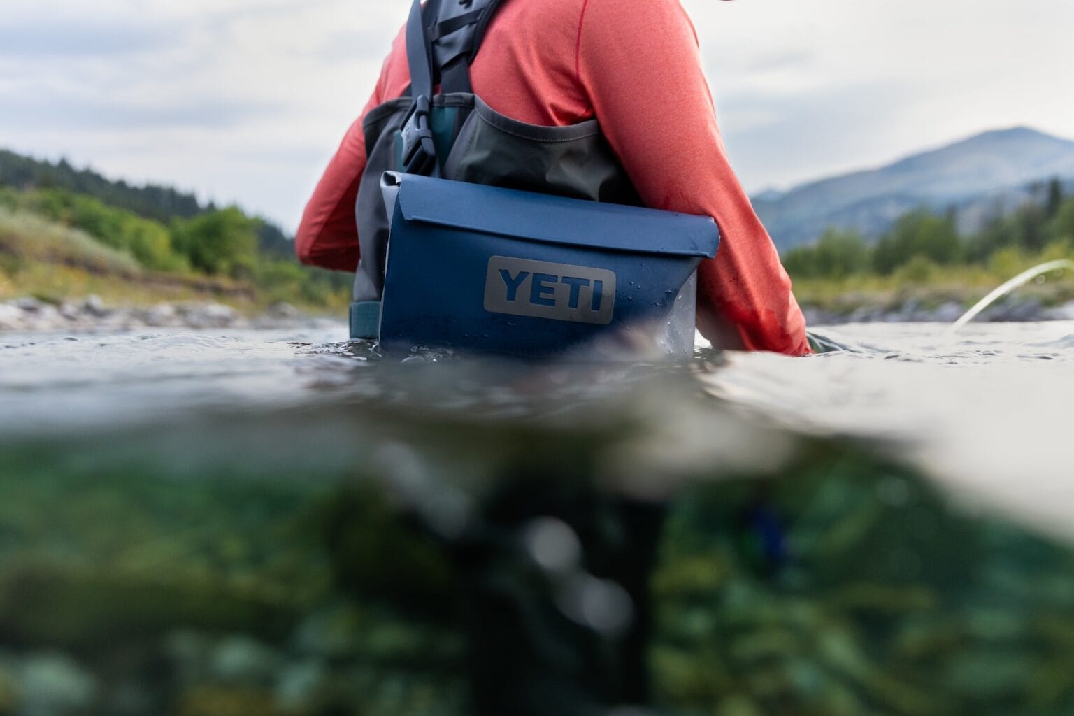 A person wades in water with a waterproof YETI bag, an ideal gift for rafters and paddlers.