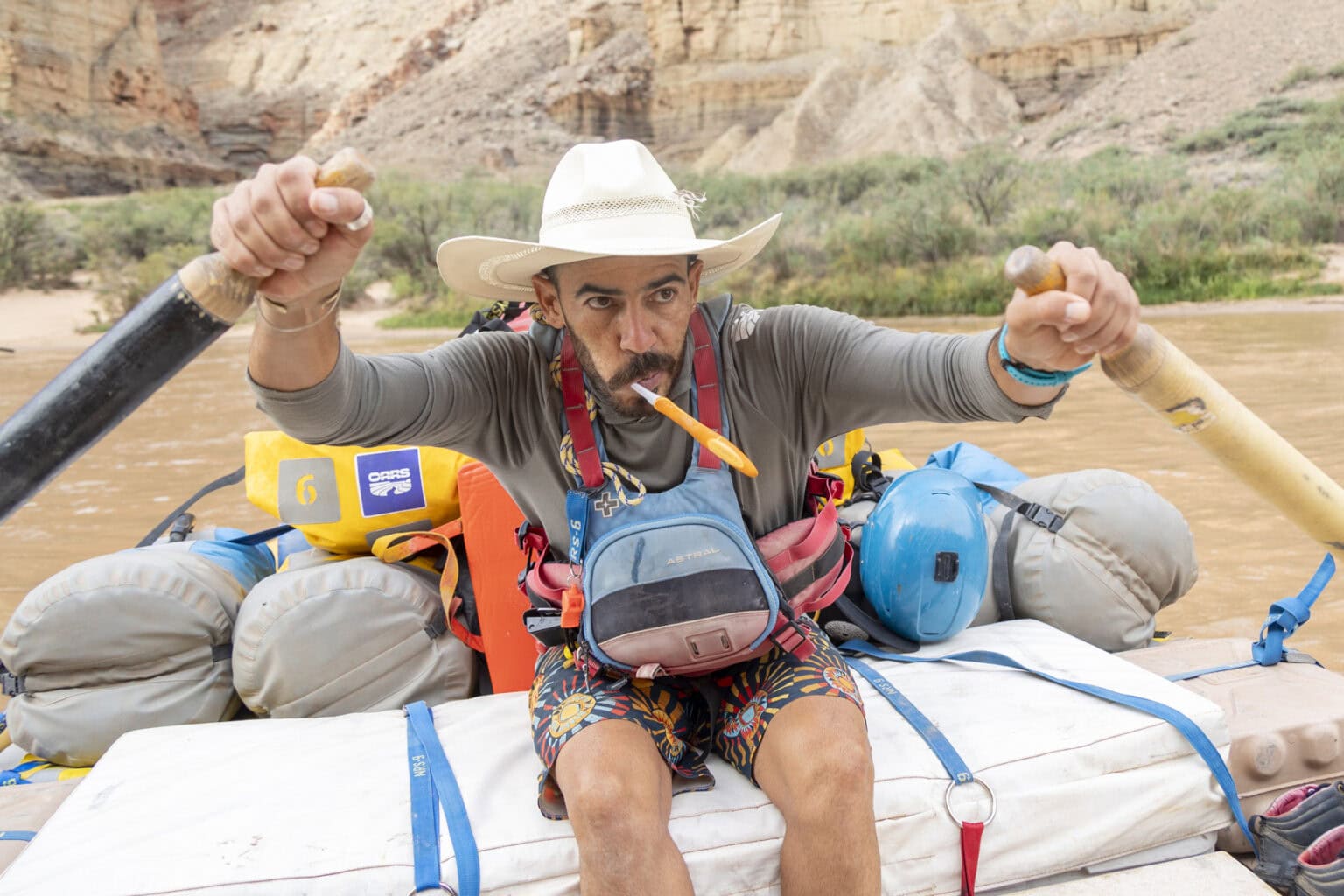 A river guide wearing a grey hoodie, rows a raft with a toothbrush in his mouth