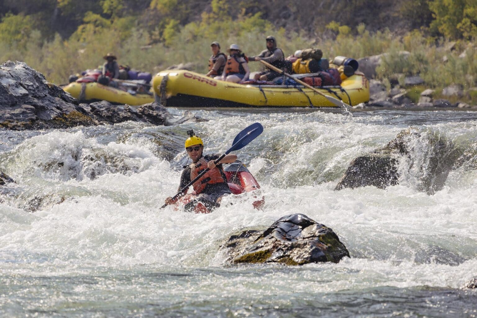 Two rafts with passengers look on as a solo kayaker challenges whitewater rapids on a river