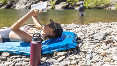 A woman reading a book and lying on her back on an inflatable blue pad situated on a rocky shore next to a river where a man is fishing in the background.