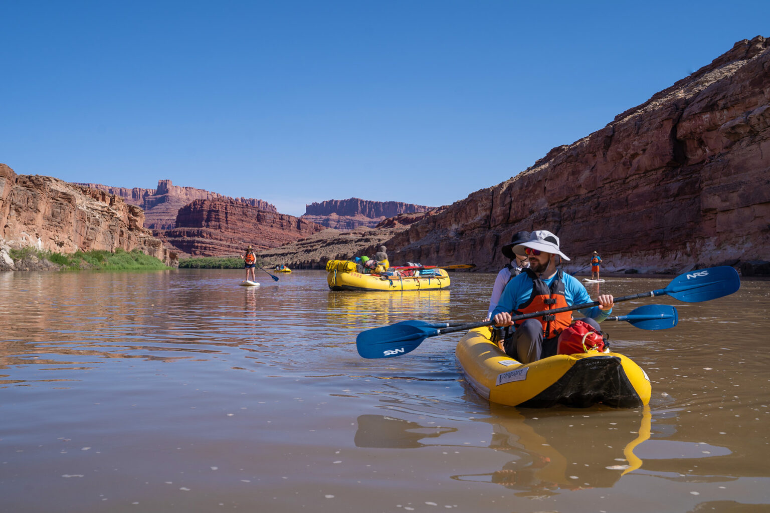 People in various watercraft on the Colorado River, including stand-up paddleboards, inflatable kayaks, and oar rafts