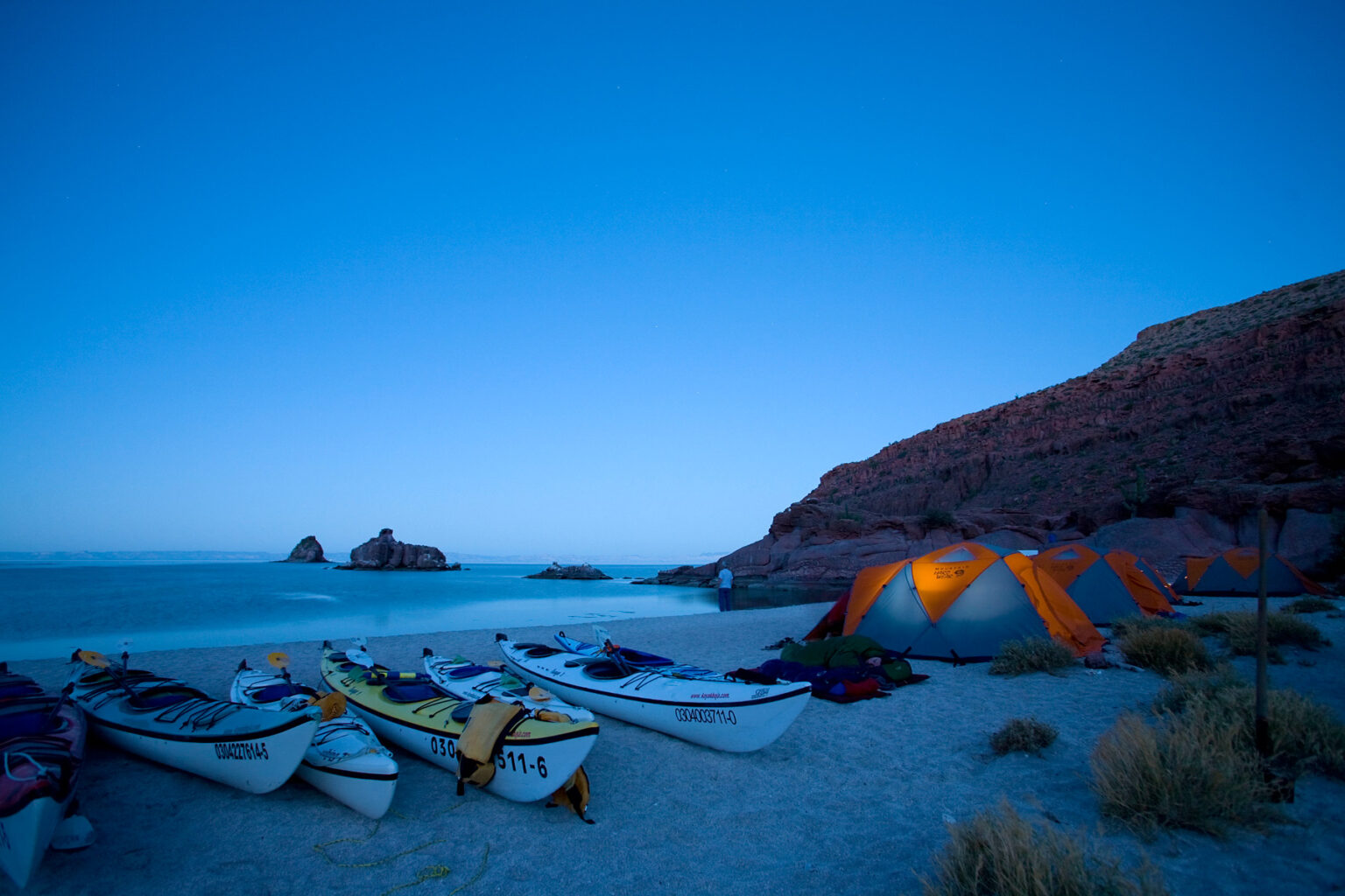 Twilight view of sea kayaks and tents on a sandy beach next to the ocean in Baja Mexico
