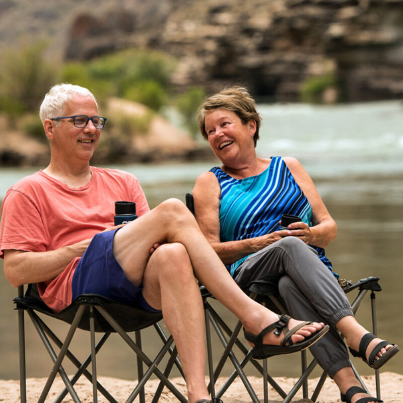 A smiling couple sits next to each other in their camp chairs on a sandy beach in Grand Canyon