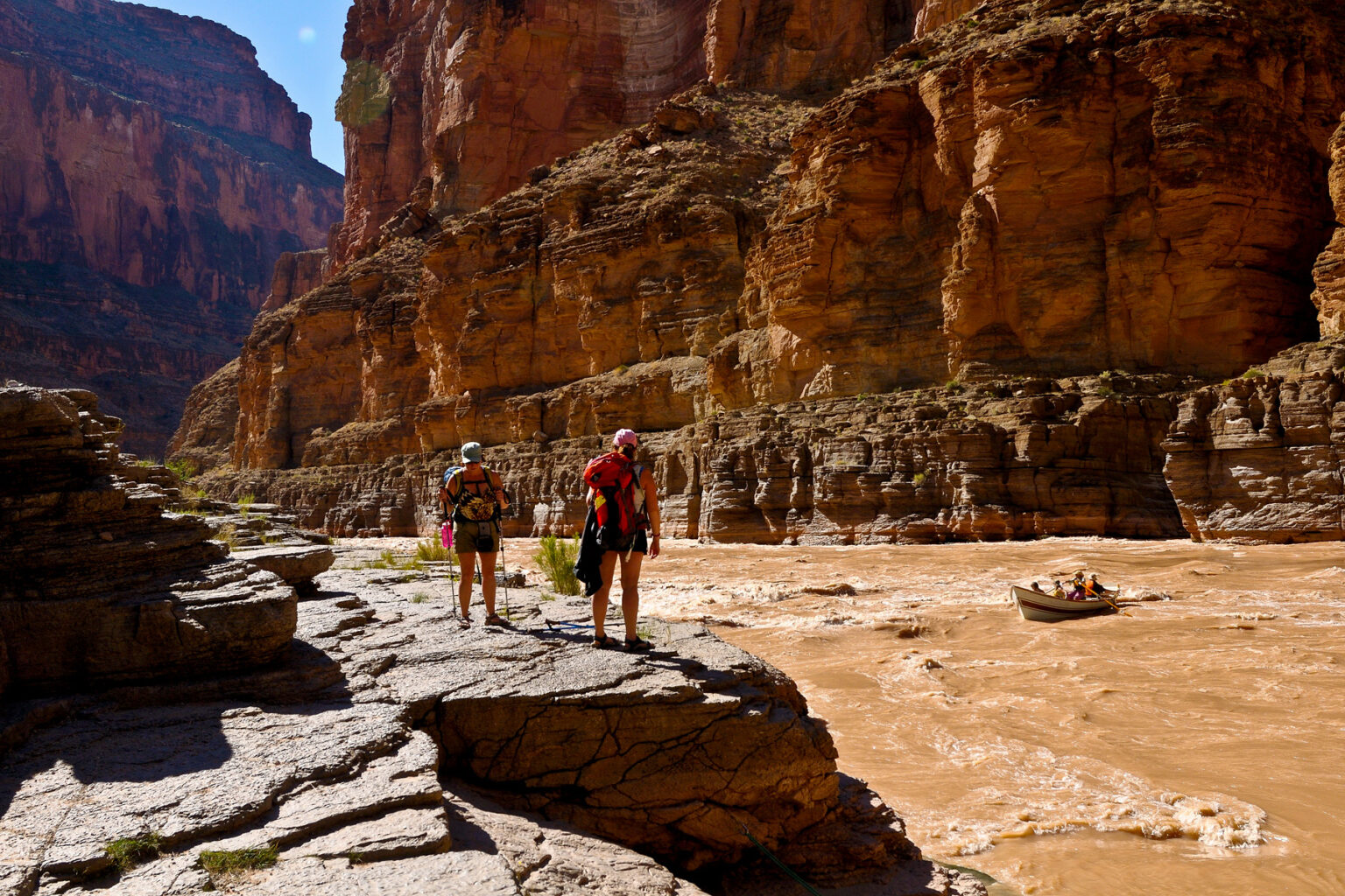 Two hikers tand on a rocky ledge and watch a dory about to enter a rapid on the Coloardo River through Grand Canyon