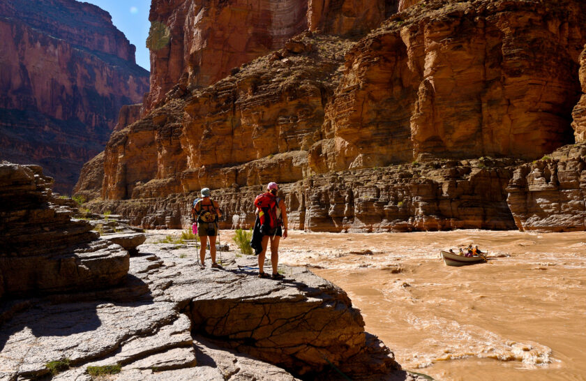 Two hikers tand on a rocky ledge and watch a dory about to enter a rapid on the Coloardo River through Grand Canyon