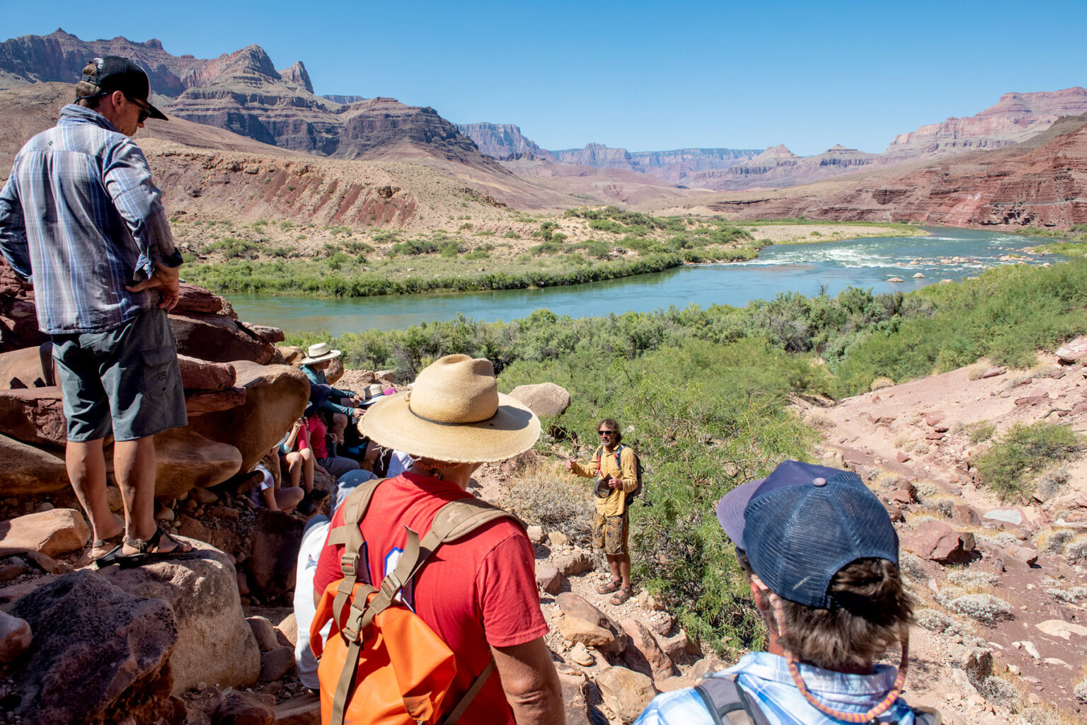 Guests sit and stand around a rocky ledge as a guide gives a talk above the banks of the Colorado River in Grand Canyon