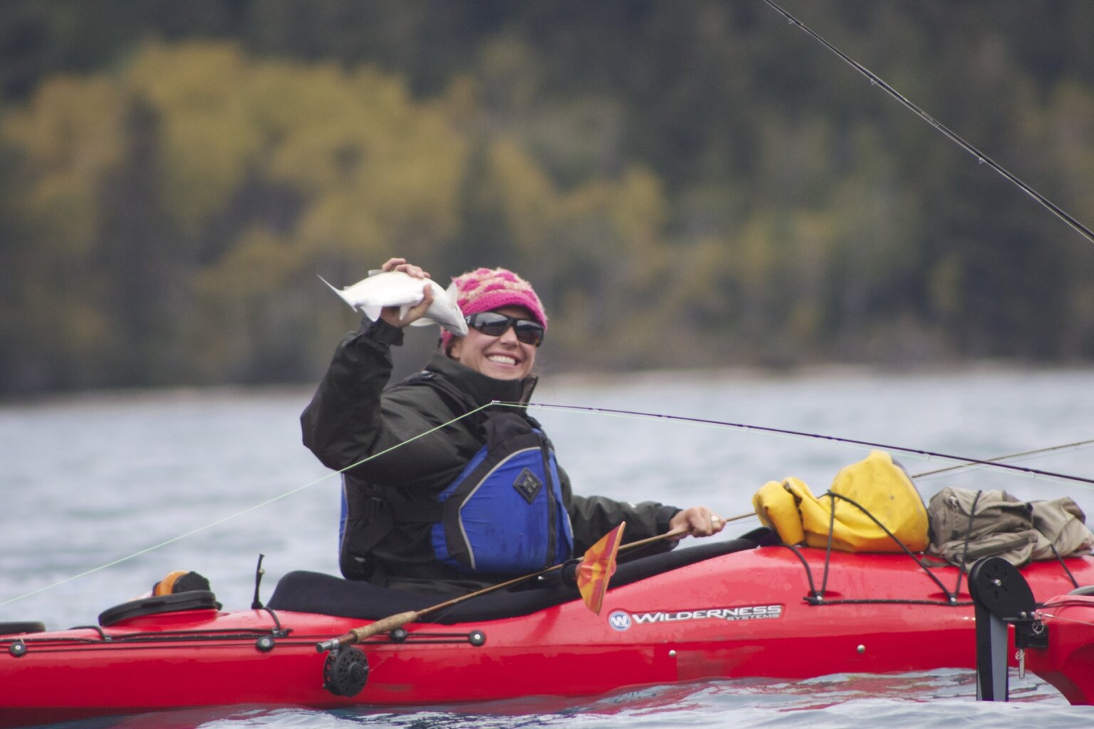 A woman in a hard shelled kayak smiles at the camera and holds up a fish she caught on Chilko Lake at Bear Camp.