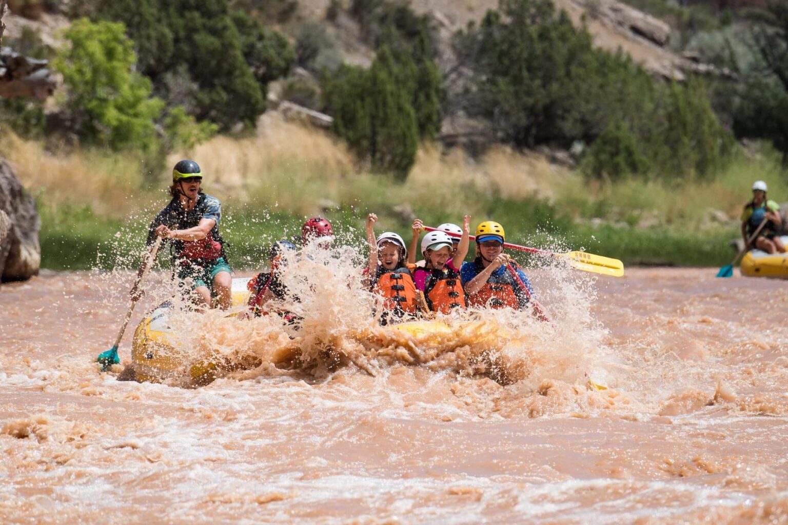 Two children with their hands in the air sit in the front of a yellow raft with adults paddling through a rapid on the Green River in Split Mountain Canyon; everyone is smiling and having a blast.