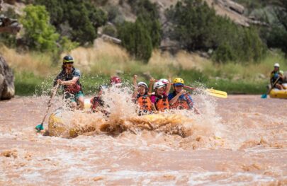 Two children with their hands in the air sit in the front of a yellow raft with adults paddling through a rapid on the Green River in Split Mountain Canyon; everyone is smiling and having a blast.