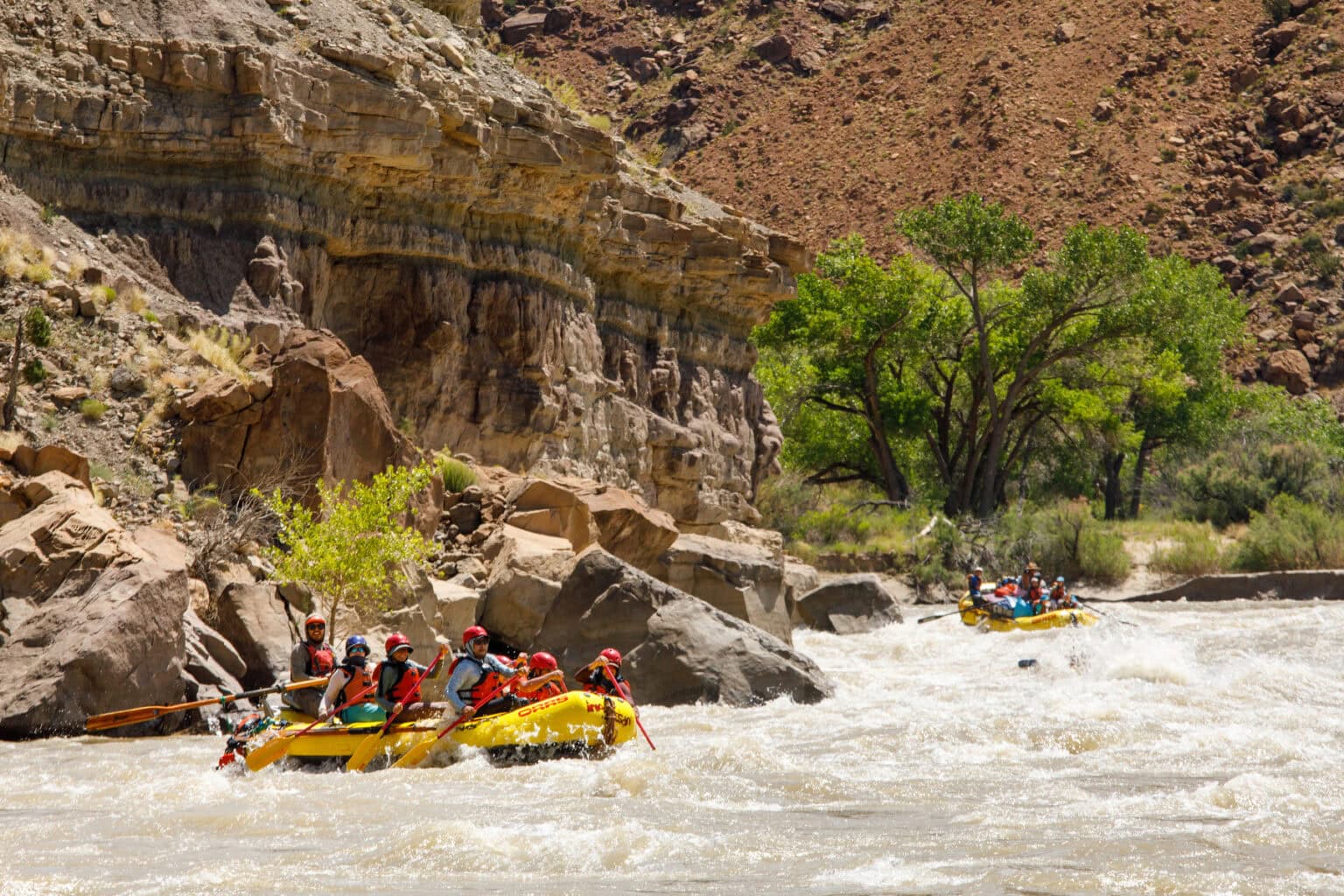 Two yellow rafts navigate a turbulent section of the Green River surrounded by rocky cliffs and greenery, under a bright sunny sky.