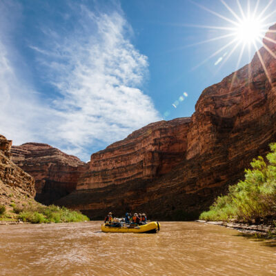 A yellow raft with people floats down the San Juan River with a sunburst above the canyon walls