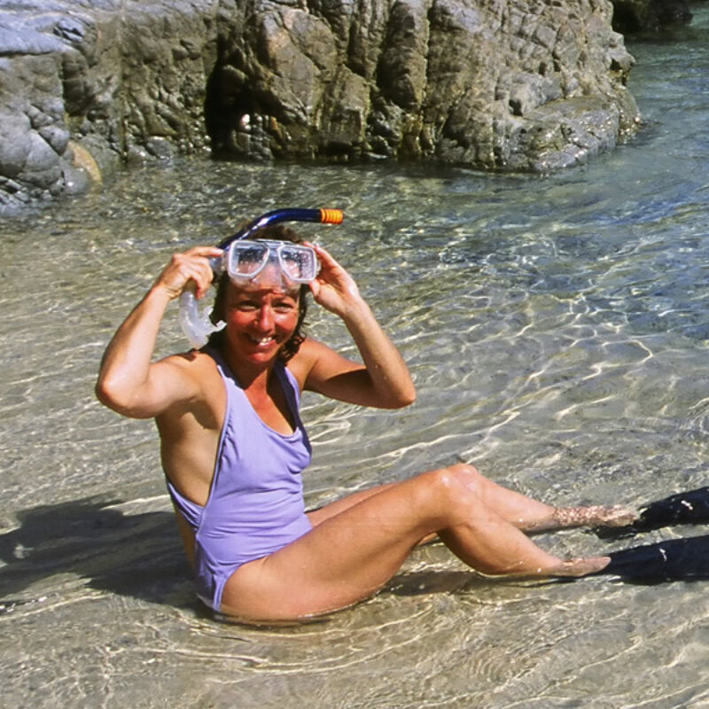 A woman in a purple bathing suit and snorkel mask sits in the water at the edge of the ocean and smiles.