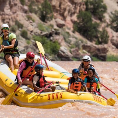 Three children sit in the front of a paddle raft with two adults and a guide behind them all smiling and having a blast on the Green River through Split Mountain Canyon