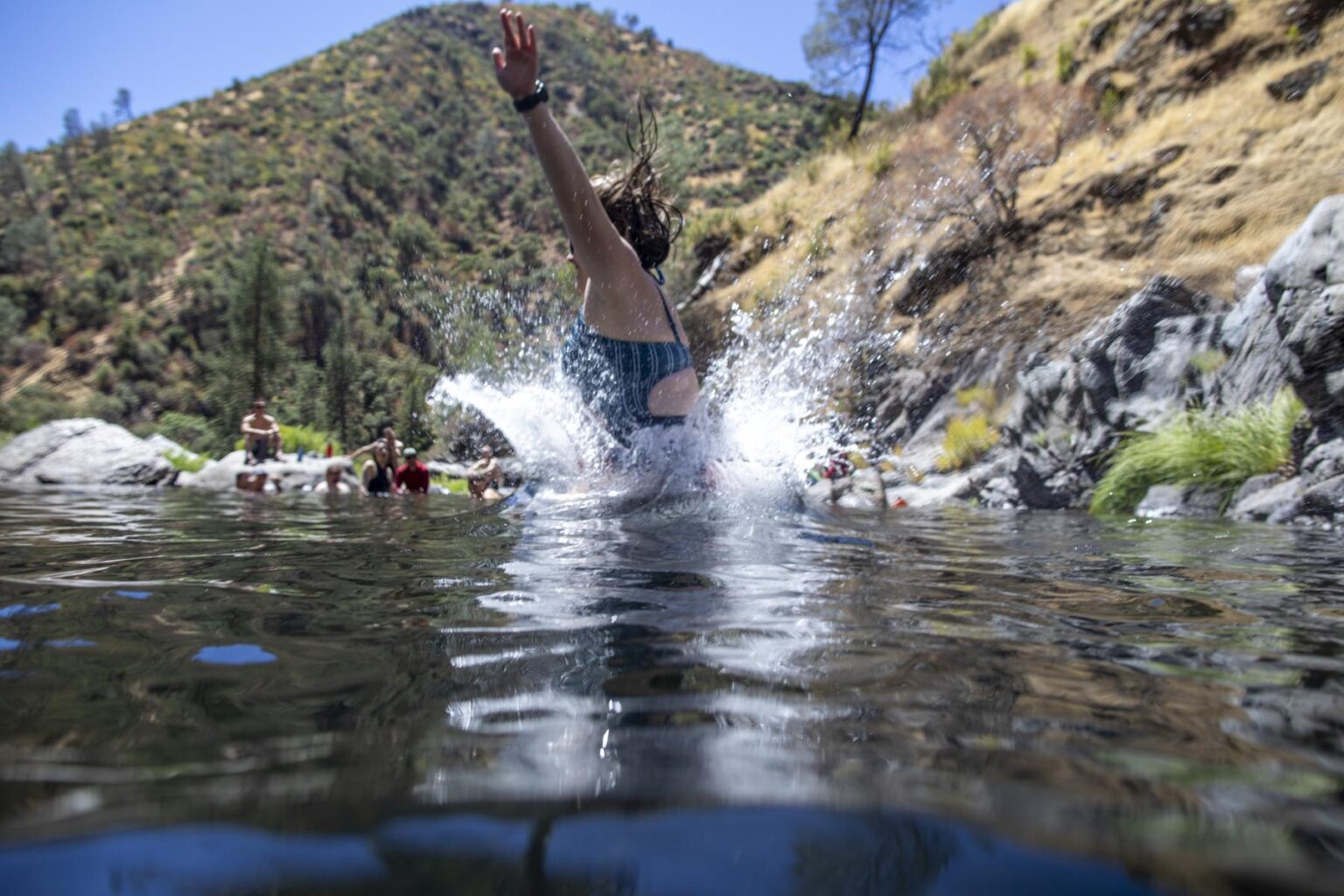 A woman jumps into a clear river with people watching in the background