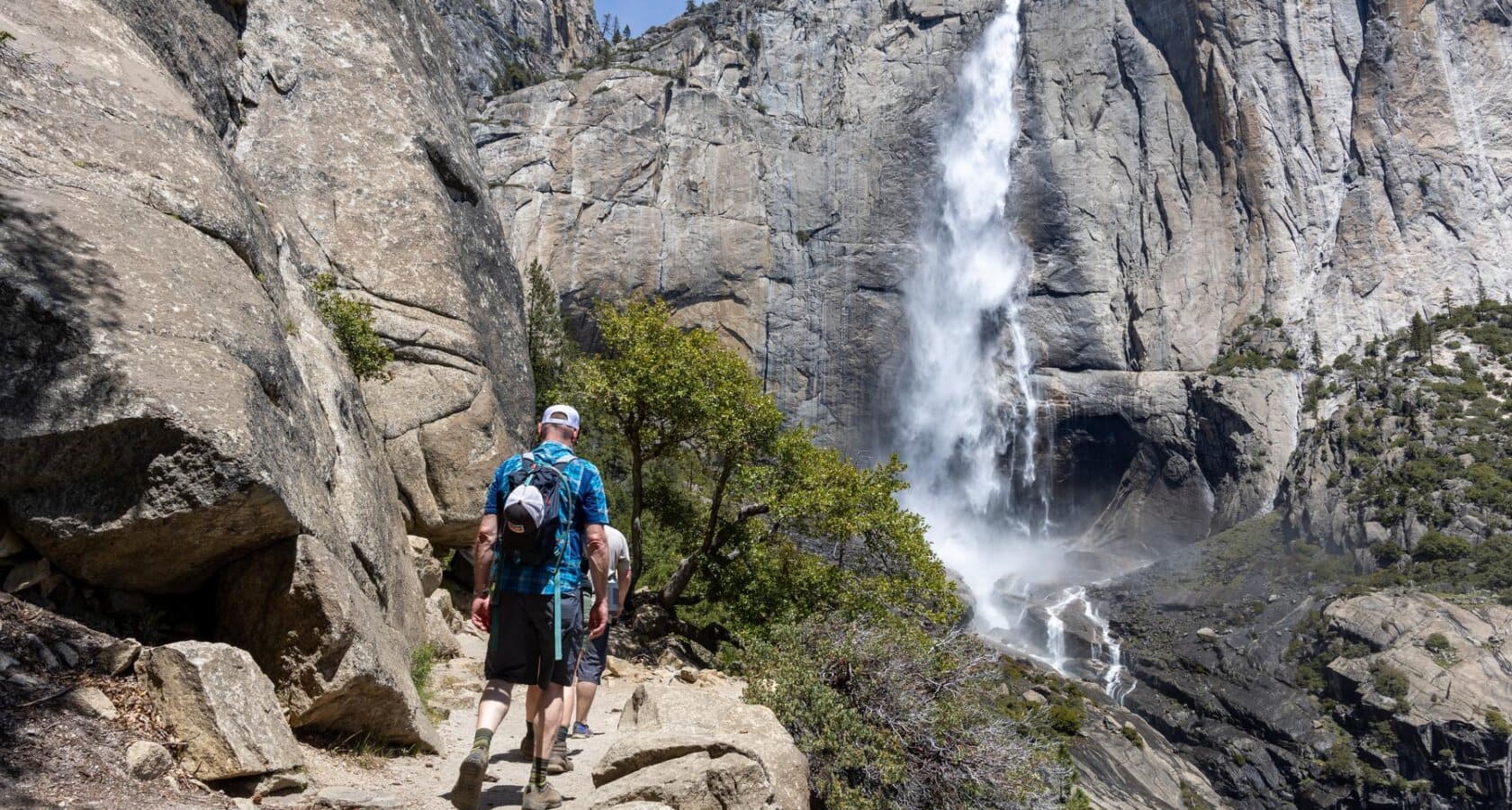 Two hikers in Yosemite National Park on a rocky trail with stunning Yosemite Falls cascading down a towering cliff in the background, surrounded by greenery.