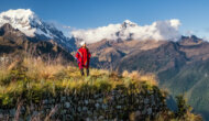 A woman in a Peruvian poncho stands on a vista overlooking forrested and snow-capped mountains.