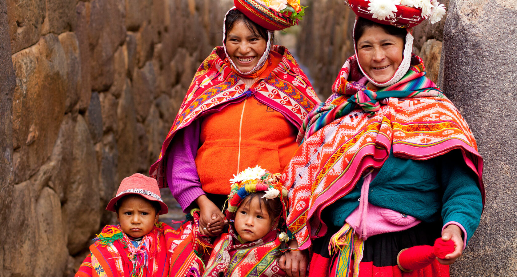 Two Peruvian women and two children adorned with Peruvian hand-crafted clothing and knit blankets pose for a photo.