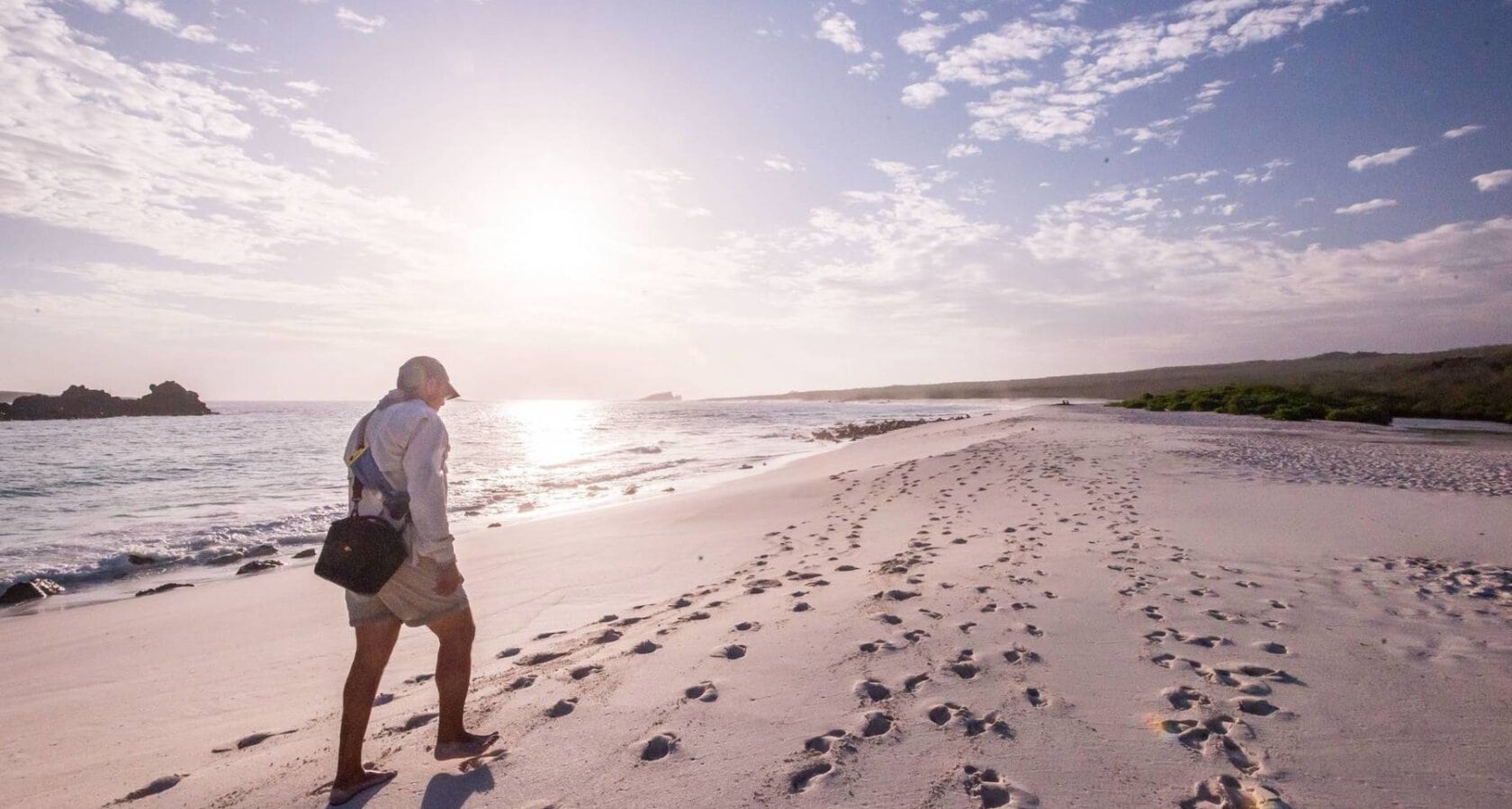 A person walks along a quiet beach in the Galapagos at sunset, leaving footprints in the soft sand, with gentle waves lapping at the shore.