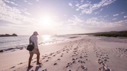 A person walks along a quiet beach in the Galapagos at sunset, leaving footprints in the soft sand, with gentle waves lapping at the shore.