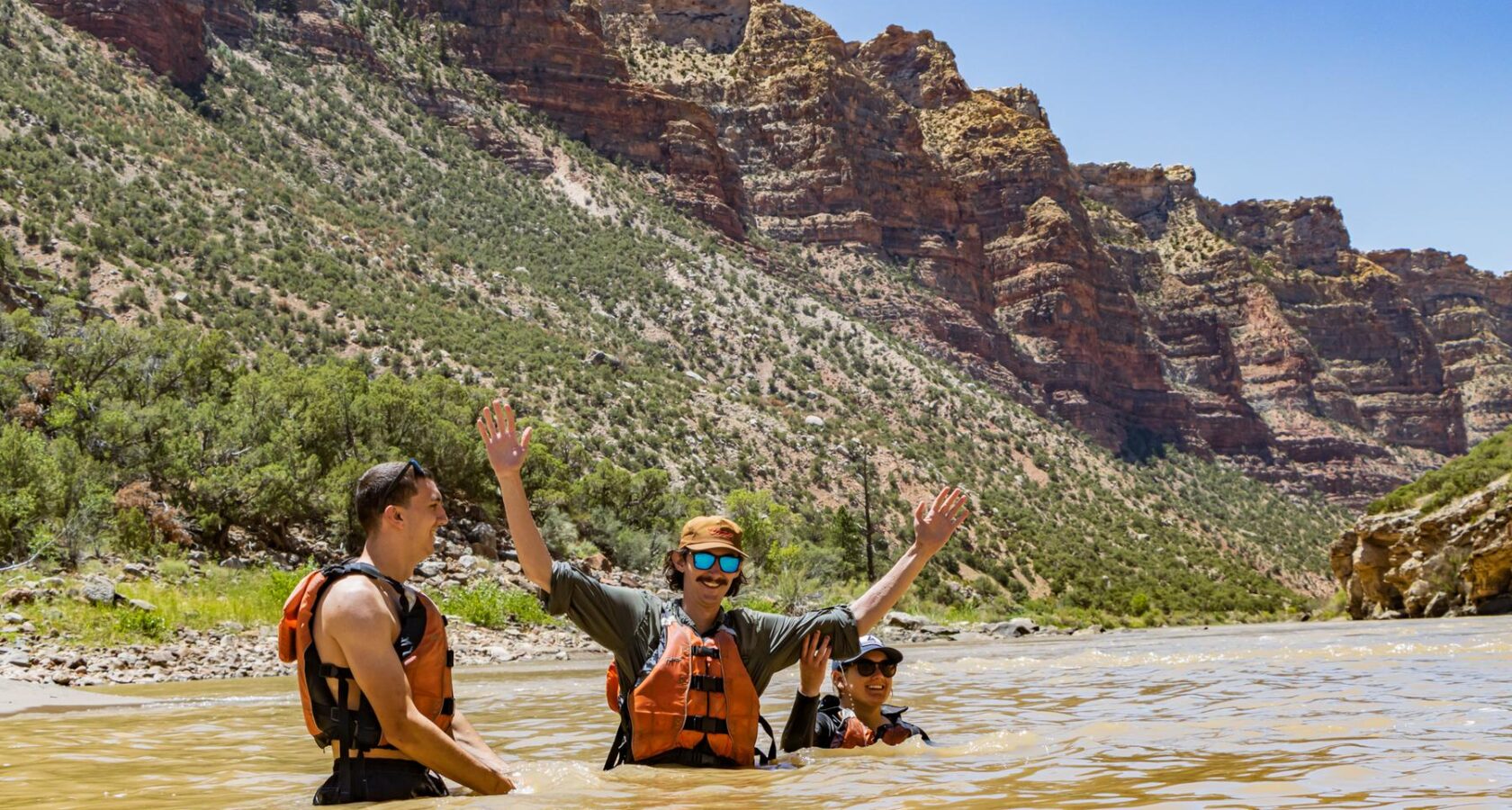3 men standing in the Green River with a sandstone canyon walls behind them
