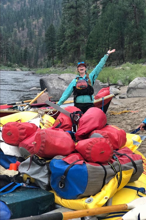 A river guide in a PFD and rain jacket stands on her raft full of gear in the rain
