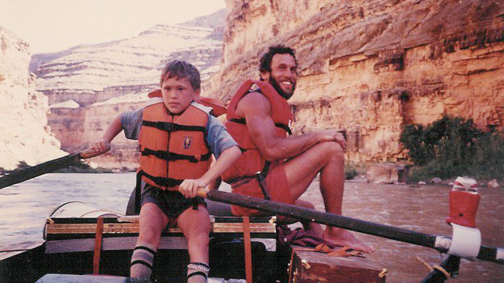 An older photograph of a father and son wearing life jackets while floating down a desert river on a raft