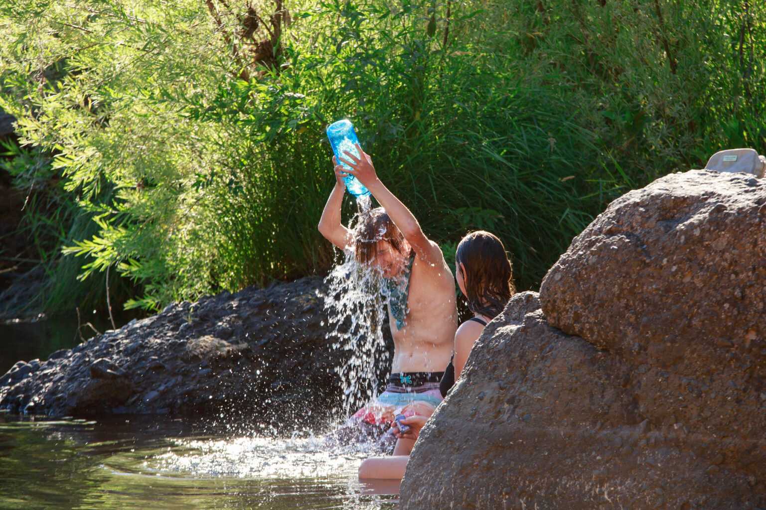 A boy kneeling in a river and using a bottle to pour water over his head