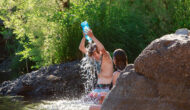 A boy kneeling in a river and using a bottle to pour water over his head