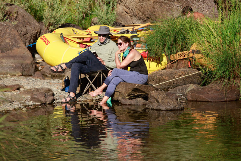 A man reading a book sits riverside with a woman whose dipping her feet in the water