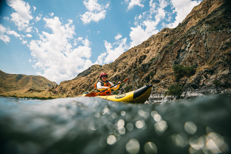 A person in a yellow inflatable kayak paddles down the Snake River with lush hills and mountains in the background.