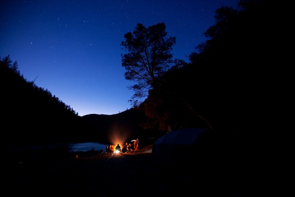 A group of people gathered around a camp fire at dusk in a scenic river canyon.