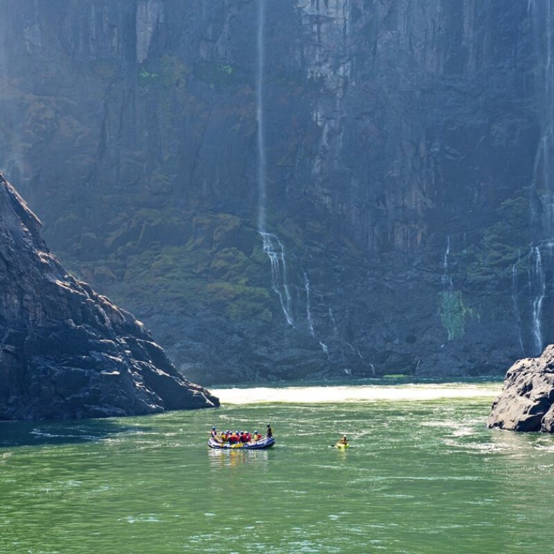 A paddle raft full of people and a person in an inflatable kayak on the Zambezi River with massive canyon walls and waterfalls stopping to admire the impressive view.