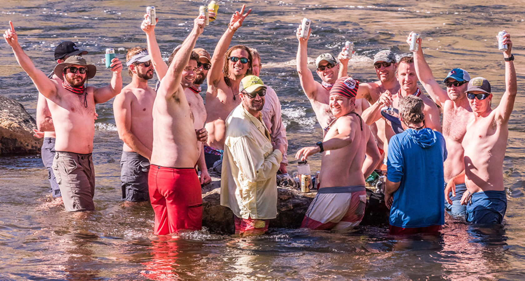 A group of men standing in the Tuolumne River smile and raise their beverages in the air while looking at the camera