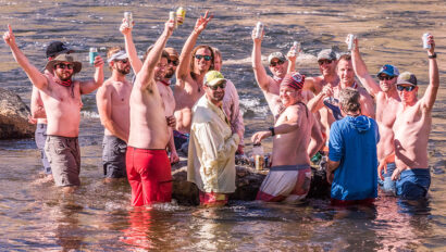 A group of men standing in the Tuolumne River smile and raise their beverages in the air while looking at the camera