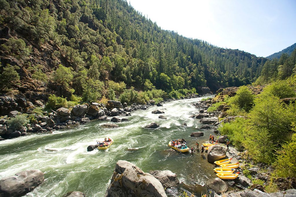 A yellow raft enters a stretch of whitewater as a second boat begins to launch and several inflatable kayaks lined up on the shore in the eddy.