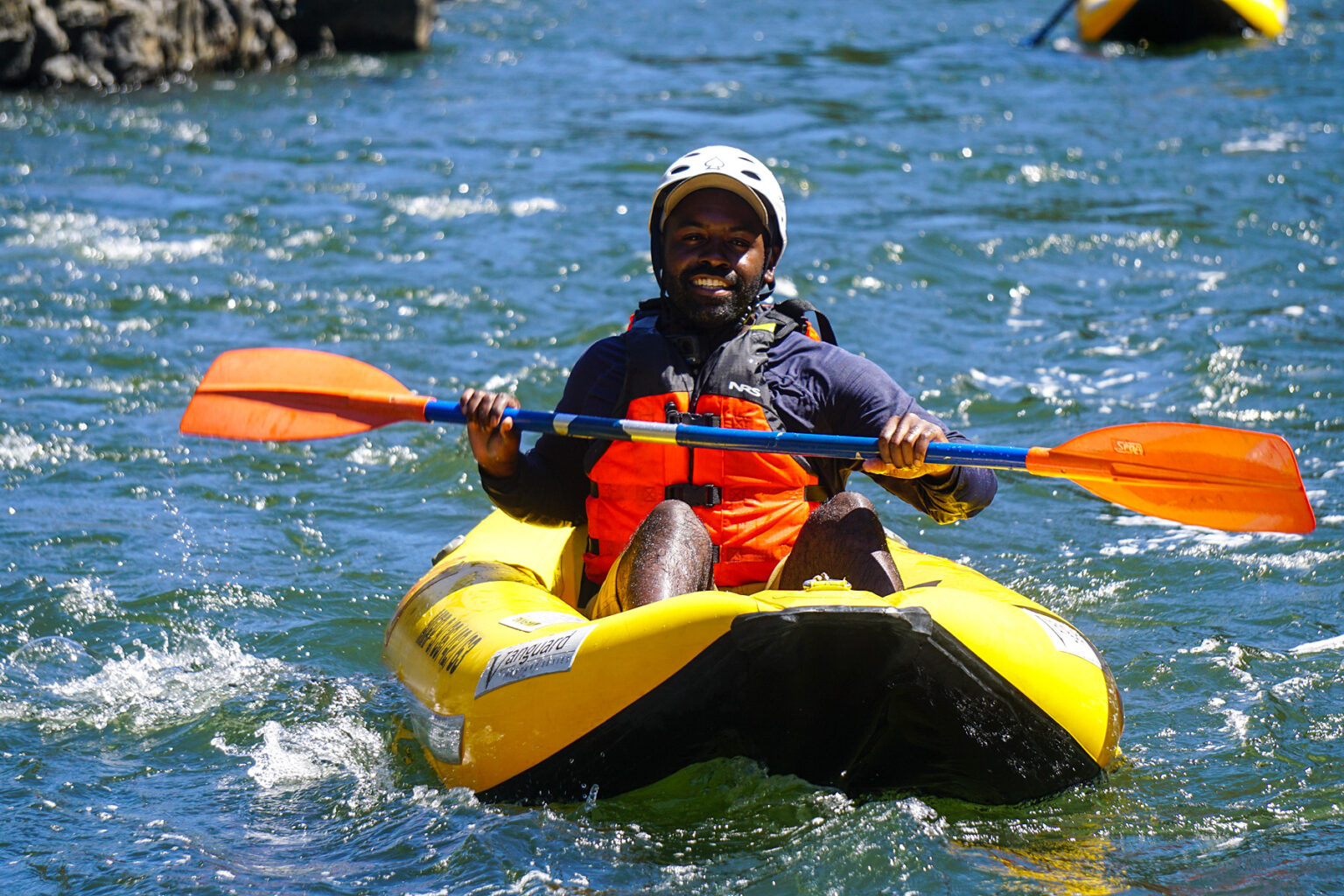 Person of color in an inflatable kayak smiles for the camera while floating down the Rogue River