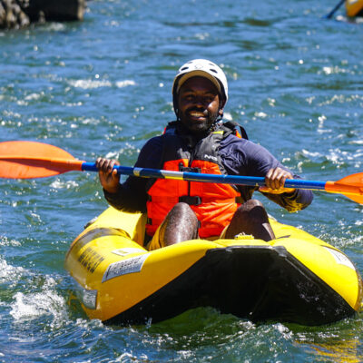 Person of color in an inflatable kayak smiles for the camera while floating down the Rogue River