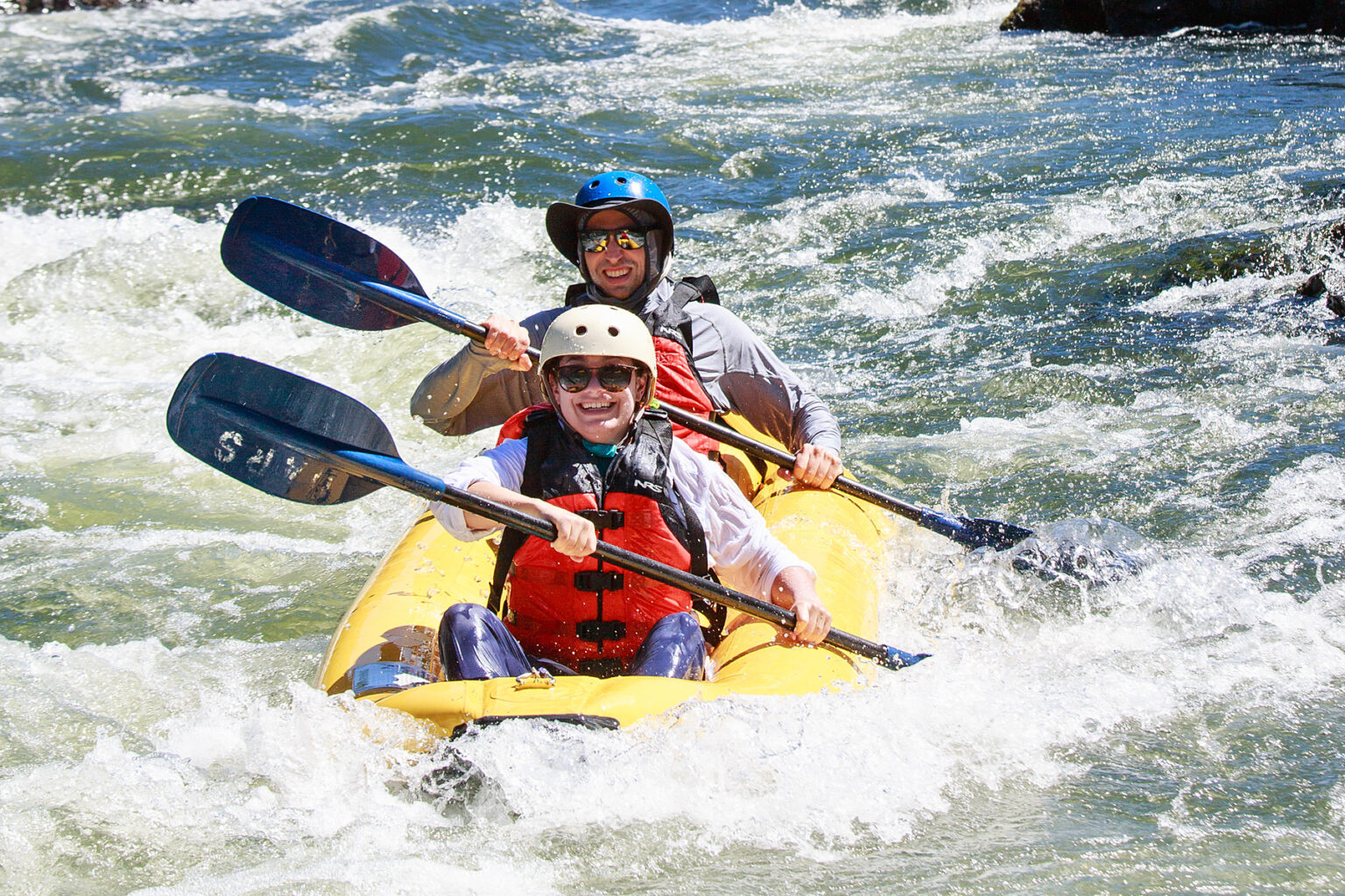 Helmeted duo smile for the camera as they paddle their yellow inflatable kayak through light rapids on the Rogue River