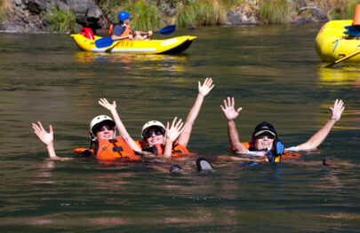 Three swimmers wave to the camera in calm waters of the Rogue River