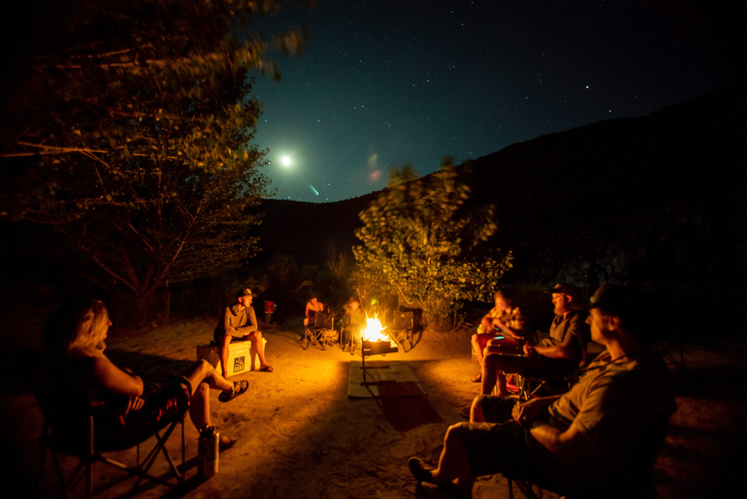 A campfire lights guides and guests as one plays the guitar. Above, the stars and moon shine brightly over Westwater Canyon, UT