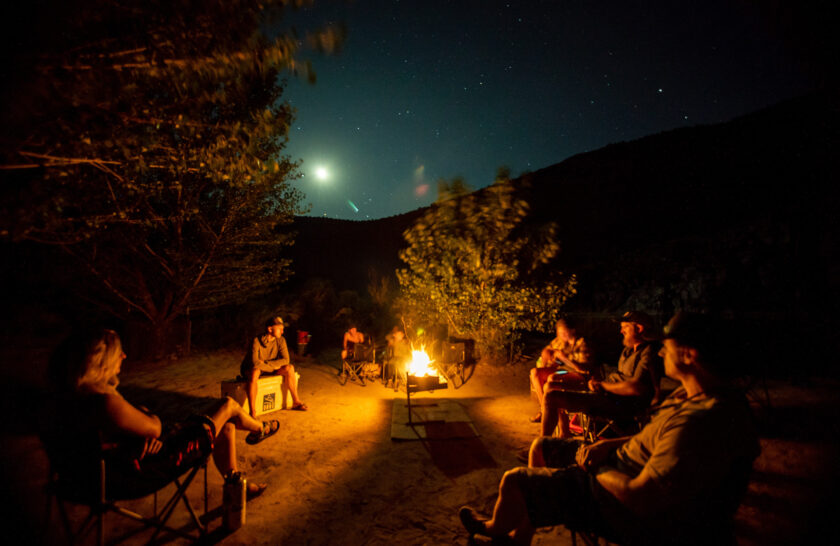 A campfire lights guides and guests as one plays the guitar. Above, the stars and moon shine brightly over Westwater Canyon, UT