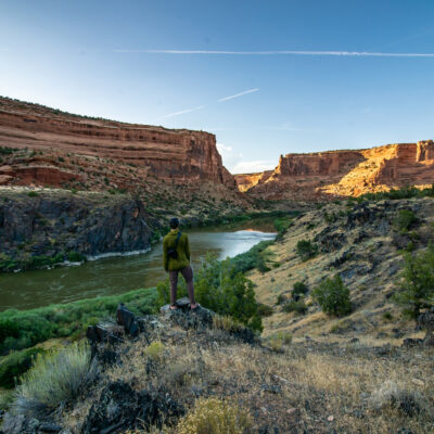 A man in a green shirt looks at the river from above as the sun lights the canyon walls of Westewater Canyon, UT