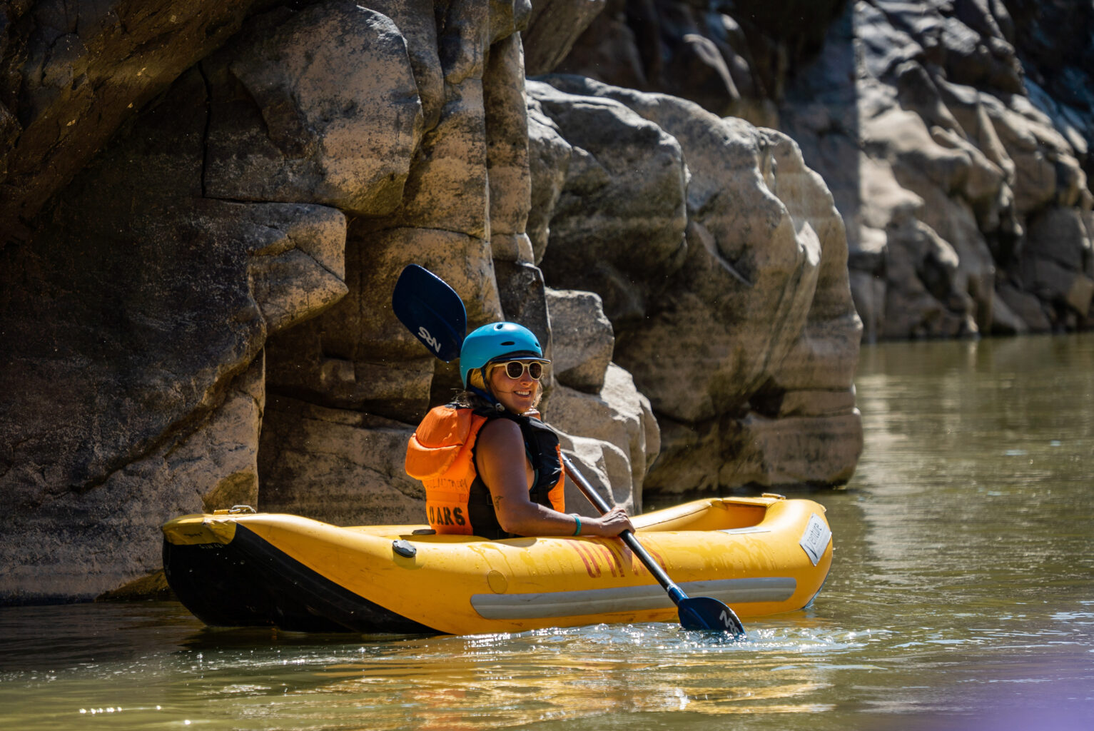 A young woman in helmet and sunglasses looks back at the camera as she paddles her inflatable kayak in calm water close to a weathered cliff face in Westwater Canyon, UT on an OARS rafting adventure