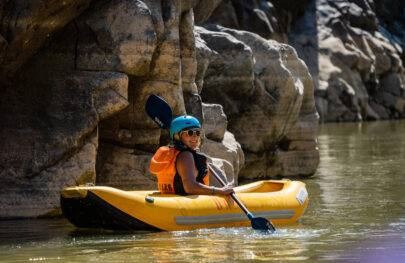 A young woman in helmet and sunglasses looks back at the camera as she paddles her inflatable kayak in calm water close to a weathered cliff face in Westwater Canyon, UT on an OARS rafting adventure
