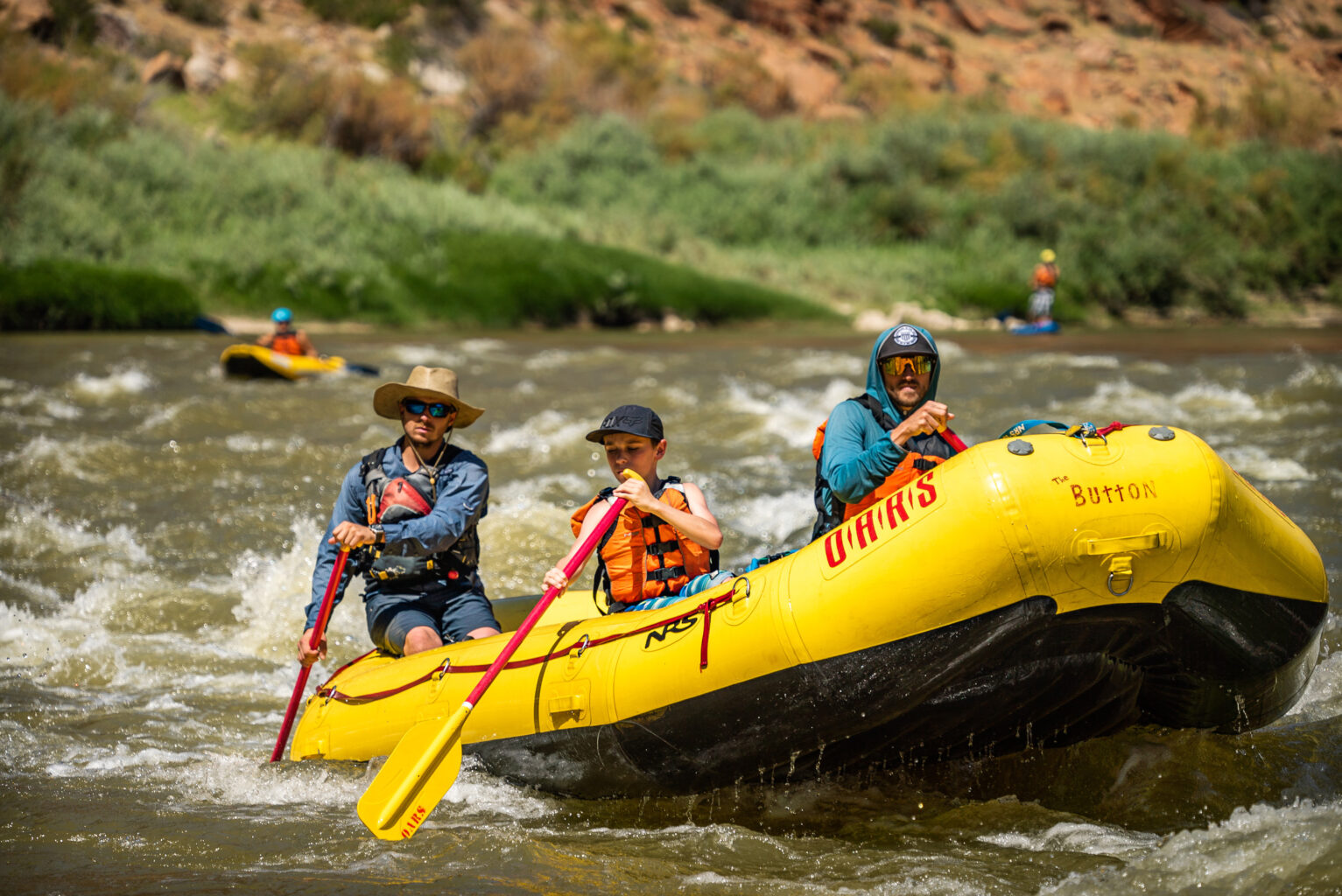 A guide in a large hat paddles two guests including a pre-teen boy in Class II whitewater on the Colorado River through Westwater Canyon, Ut. In the background, a person in an inflatable kayak and another on a stand-up paddleboat tackle the fun waves on this OARS rafting adventure