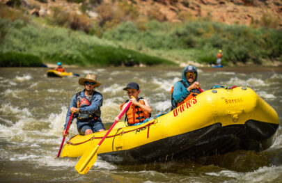 A guide in a large hat paddles two guests including a pre-teen boy in Class II whitewater on the Colorado River through Westwater Canyon, Ut. In the background, a person in an inflatable kayak and another on a stand-up paddleboat tackle the fun waves on this OARS rafting adventure