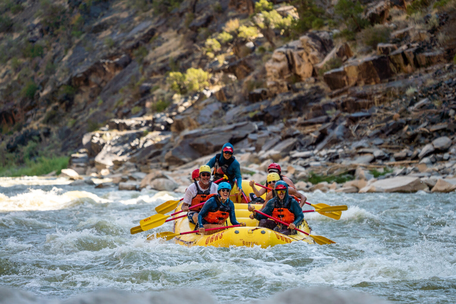 Six guests and a guide in a paddle raft laughing while enjoying Class II rapids on an OARS trip in Westwater Canyon, UT