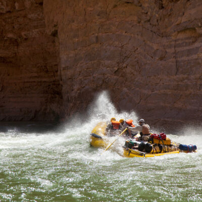 OARS raft hits rapid sending spray up in contrast to dark canyon walls
