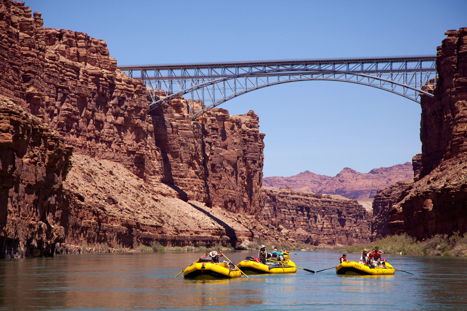 Three yellow OARS rafts travel under bridge near put in at Lees Ferry in Grand Canyon