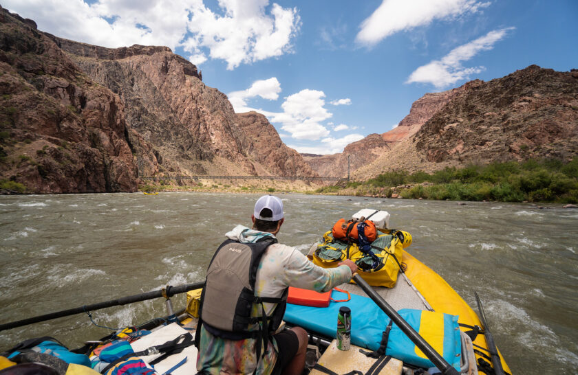 OARS rafting guide steers his raft down the Colorado River with a foot bridge to Phantom Ranch in the distance